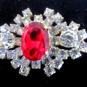 May include: An ornate vintage brooch featuring a large, oval, ruby-red central stone, encircled by numerous clear, sparkling rhinestones. The brooch has a gold-tone metal setting, set against a black backdrop. The design is intricate and glamorous.