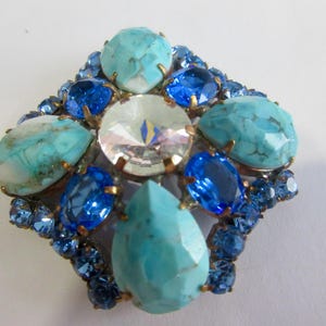 Peut inclure: Broche vintage ornée d'une pierre centrale transparente et facettée, entourée de pierres bleues et turquoise. La broche est en forme de losange, avec de petites pierres bleues sur le pourtour. Les pierres sont serties dans un métal doré.