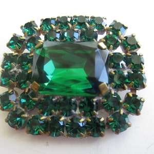 May include: A vintage green rhinestone brooch with a large emerald green center stone. The brooch is made of metal and has a gold tone finish.