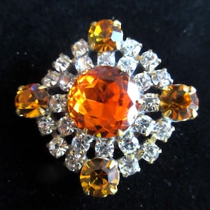 May include: A large orange rhinestone surrounded by smaller clear rhinestones in a square shape.