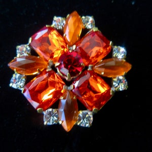 May include: A vintage brooch with a cluster of orange and red rhinestones set in gold tone metal. The brooch has a starburst design with clear rhinestones around the edges.