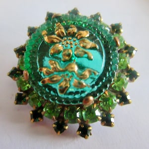 May include: A vintage brooch with a central teal-colored glass circle featuring a gold floral design. The brooch is surrounded by green and dark green rhinestones, creating a starburst effect. The metal has a gold tone.
