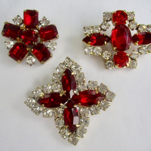 May include: Three red rhinestone brooches with clear rhinestone accents. The brooches are in the shape of a flower, a cross, and a star.