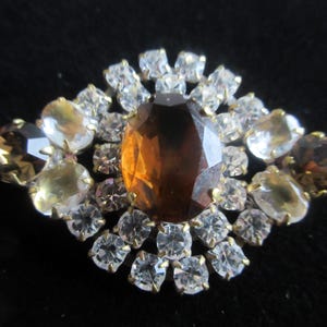 May include: A vintage brooch featuring a large, faceted, amber-colored central stone surrounded by smaller clear and amber-toned crystals. The brooch has a gold-tone metal setting and a symmetrical, floral-like design. The crystals are round and oval-shaped.