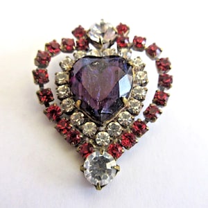 May include: Vintage heart-shaped brooch with a large, faceted purple center stone. The brooch is surrounded by clear and red rhinestones, set in a gold-tone metal frame. A classic piece of jewelry.