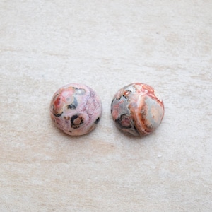 May include: Two circular, polished cabochons featuring intricate patterns of pink, red, white, gray, and black. The left stone displays a soft pink base with dark circular inclusions, while the right stone exhibits a marbled blend of red, white, and gray tones.
