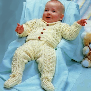 May include: A baby wearing a crocheted cream-colored sweater and pants set. The outfit features a cable knit pattern and button closure.