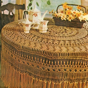 May include: A brown crocheted tablecloth with a fringe border. The tablecloth is laid on a round table with a white teapot and creamer set in the center.