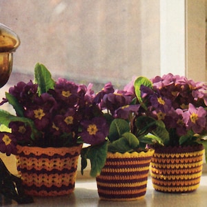 May include: Three purple and yellow crocheted plant pots with purple flowers in them. The pots are sitting on a windowsill.