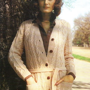 May include: A light beige cable-knit cardigan with a matching belt and large brown buttons. The cardigan has pockets and is worn over a brown collared shirt. The woman is standing next to a tree.