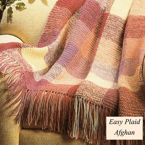 May include: A crocheted plaid throw blanket in shades of pink, purple, and cream with a fringe border. The blanket is draped over a tan armchair. The text "Easy Plaid Afghan" is printed on the image.