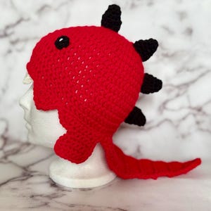 Könnte beinhalten: Ein roter gehäkelter Dinosaurierhut mit schwarzen Stacheln und einem Schwanz. Der Hut hat ein schwarzes Auge und weiße Zähne, was ein verspieltes Design ergibt. Dieser handgefertigte Hut ist perfekt für Kostümpartys oder den Alltag.