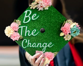 May include: Green graduation cap with white glitter and the words "Be the Change" in white lettering. The cap is decorated with pink, white, and blue flowers.