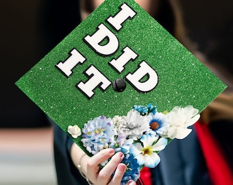 May include: Green graduation cap with glitter and the words "I DID IT" in white letters. The cap is decorated with blue and white flowers.