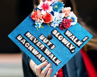 May include: Blue graduation cap with white glitter lettering that reads "Walk Slowly, But I Never Walk Backward". The cap is decorated with a variety of colorful flowers.