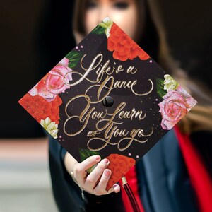 May include: Black graduation cap with a floral design and gold glitter. The cap has the text "Life is a dance you learn as you go" written on it.