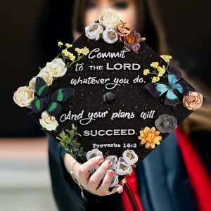 May include: Black graduation cap decorated with white and green butterflies, white and yellow flowers, and the text "Commit to the Lord whatever you do, And your plans will succeed. Proverbs 16:3"