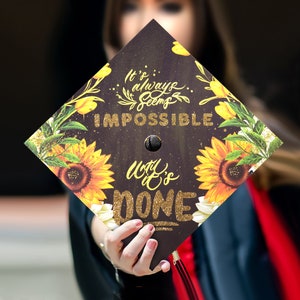 May include: A black graduation cap with a gold glitter design. The cap features the text "It's always seems impossible until it's done." with sunflowers and other floral designs.