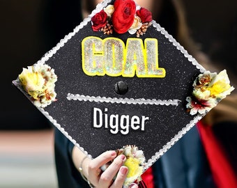 May include: Black graduation cap with white lace trim and a yellow and red floral design. The cap has the word "GOAL" in yellow glitter letters and the word "Digger" in white letters.