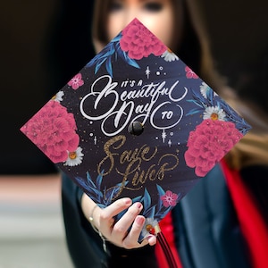 May include: A black graduation cap decorated with pink flowers and gold glitter. The cap has the text "It's a beautiful day to save lives" written on it.