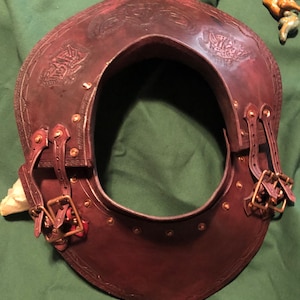 Può includere: Un collare di protezione per il collo in pelle marrone con dettagli decorativi e fibbie in ottone. Il collare è progettato per proteggere il collo e le spalle.