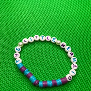 May include: White beaded bracelet with the words "The Little Mermaid" spelled out in colorful letters. The beads are separated by small purple and blue beads.