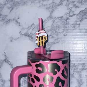 Puede incluir: Vaso rosa y morado con diseño de estampado de leopardo y asa rosa. El vaso tiene una pajita rosa con un adorno decorativo con un gorro a rayas y un emblema dorado.