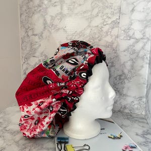 May include: A red, white, and black satin bonnet featuring the University of Georgia Bulldogs logo and text. The bonnet has a black ruffled trim and is displayed on a white mannequin head.