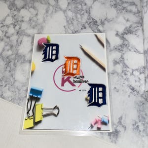 May include: A clear plastic sleeve containing patches with the Detroit Tigers logo in navy blue and orange. The patches are on a white background with the text "K Zen Kreations" and "2016". Also included are binder clips and push pins.