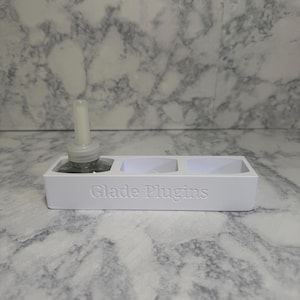 May include: A white rectangular holder with three circular cutouts designed for air freshener refills. The front of the holder features the embossed text Glade Plugins. One clear glass air freshener refill bottle is inserted into the first slot.