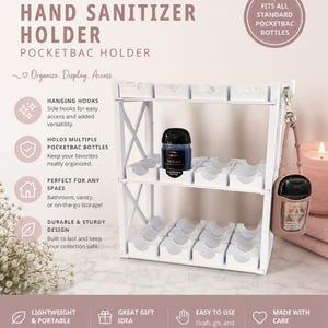 May include: A white two-tiered plastic organizer stand designed for hand sanitizer bottles. The unit features multiple molded slots for storage, side hooks for hanging accessories, and text labels detailing its use for Bath & Body Works PocketBac bottles.