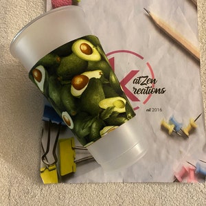 May include: A white plastic cup with a green avocado print sleeve. The sleeve has a pattern of avocados with green leaves. The cup is sitting on a white surface with a pink eraser, a pencil, and a piece of paper with the text "atZen creations est 2016".