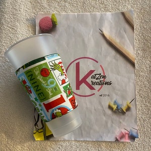 May include: A white plastic cup with a colorful fabric sleeve featuring a Grinch design. The sleeve has the text "The Grinch is Back" and "Since 1957". The cup is sitting on a sheet of paper with the text "K at Zen Creations est 2016".