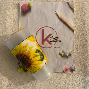 May include: A clear plastic cup with a sunflower fabric sleeve. The sleeve has a yellow sunflower with green leaves on a white background. The cup is sitting on a white towel. The background is a white sheet of paper with a pink eraser, pencils, and push pins.