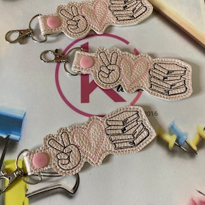 May include: Three pink leather keychains with a peace sign, a heart, and a stack of books. The keychains are embroidered with black thread and have a pink button on the top.