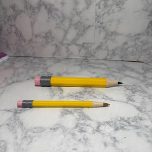 May include: Two yellow pencils with grey ferrules and pink erasers. The larger pencil has a dark graphite tip, while the smaller pencil has a brown tip. Both pencils feature a hexagonal barrel design and a light wood-colored sharpened point.