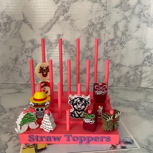 May include: A pink straw topper display stand with various decorative toppers. The toppers include a reindeer, a character with a yellow hat, a Christmas tree, a Highland cow, and Dr. Pepper and Coke bottles. The text "Straw Toppers" is visible.
