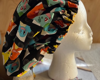 May include: A black satin hair bonnet featuring a repeating pattern of colorful cartoon characters including Pikachu, Charmander, Squirtle, and Bulbasaur. The bonnet has a gathered elastic edge and is displayed on a white foam mannequin head.
