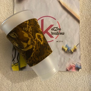 May include: A white plastic cup with a brown and black leopard print fabric sleeve. The sleeve has a close-up image of a leopard's face. The cup is sitting on a white piece of paper with the text "at Zen creations est 2016".