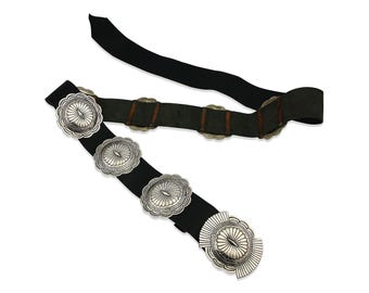 May include: Two belts are displayed against a white background. One is black with large, silver-colored, scalloped conchos. The other is a dark gray suede with smaller, silver-colored conchos and brown leather accents. Both belts have a classic, southwestern style.