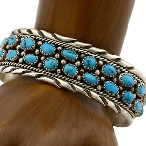 Peut inclure: Bracelet manchette en argent orné de multiples pierres turquoises ovales. Le bracelet présente un design détaillé avec une rangée de pierres turquoises serties dans une bande d'argent. Le bracelet est porté au poignet.