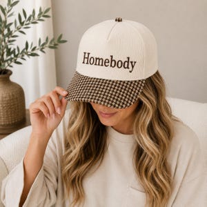 Peut inclure: Une casquette de baseball en velours côtelé crème avec le mot Homebody brodé en fil marron sur le panneau avant. La visière de la casquette présente un motif pied-de-poule marron et crème. La casquette possède une calotte structurée et un bouton.