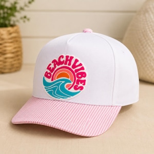 Peut inclure: Une casquette de baseball blanche avec un écusson circulaire sur le devant portant le texte BEACH VIBES en rose, entourant un graphique d'un soleil orange et rouge au-dessus d'une vague turquoise. La visière est rayée rose et blanc.