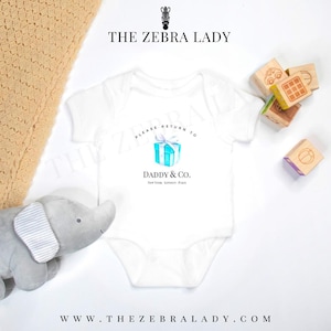 Op de afbeelding: Witte babybody met de tekst "PLEASE RETURN TO DADDY & CO." en een blauwe cadeauverpakking. De afbeelding bevat een grijze olifant, houten blokken en een beige deken. Het logo van The Zebra Lady is zichtbaar.