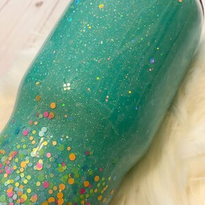 May include: A teal green tumbler with a glitter finish and a confetti design. The confetti is in various colors, including pink, yellow, green, and blue.