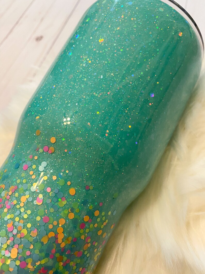 May include: A teal green tumbler with a glitter finish and a confetti design. The confetti is in various colors, including pink, yellow, green, and blue.