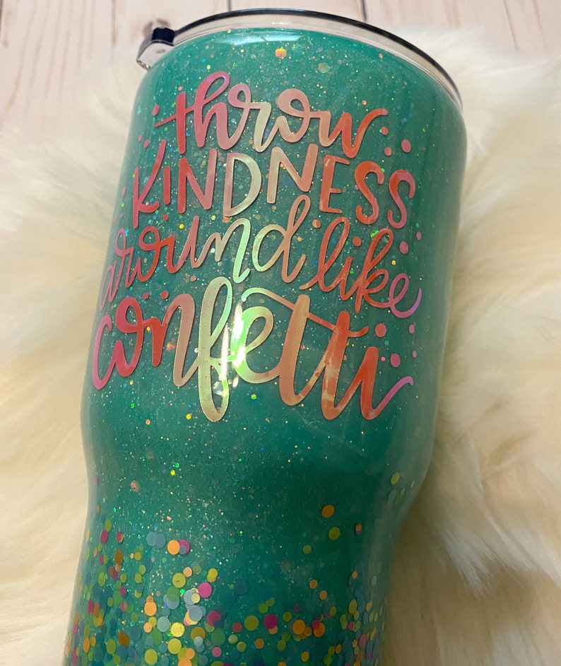 May include: A teal green tumbler with glitter and confetti. The tumbler has the text "Throw kindness around like confetti" in a pink and orange gradient.