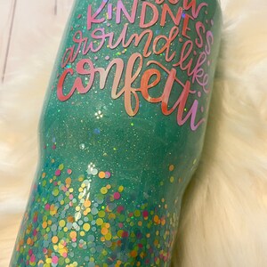 May include: A teal green tumbler with a glittery finish and colorful confetti at the bottom. The tumbler has the text "Throw Kindness Around Like Confetti" in a rainbow gradient.
