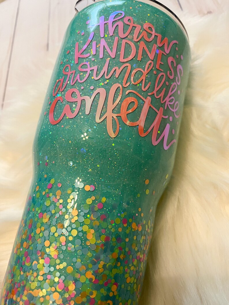 May include: A teal green tumbler with a glittery finish and colorful confetti at the bottom. The tumbler has the text "Throw Kindness Around Like Confetti" in a rainbow gradient.