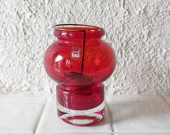 May include: A red glass vase with a clear glass base. The vase has a rounded body and a small opening at the top. The vase is sitting on a white tiled surface.